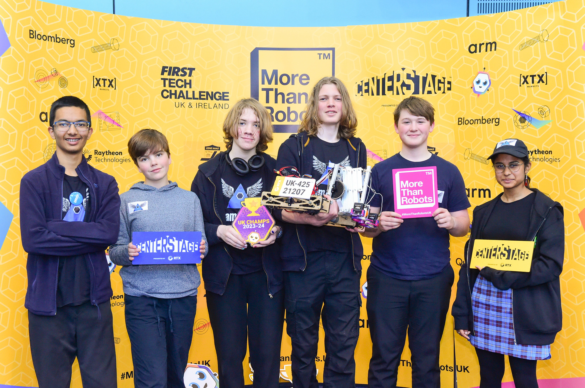 Five people standing in front of a yellow backdrop. The middle holds a robot.